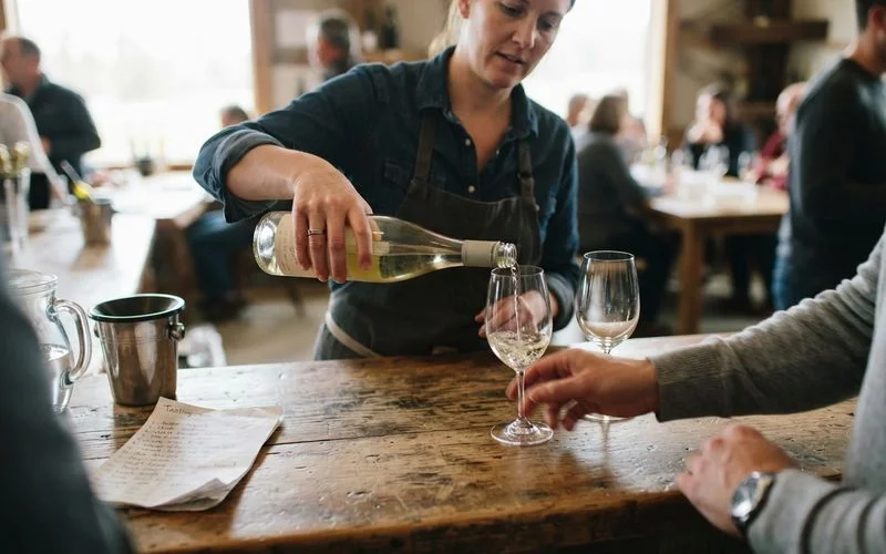 A host pouring a small measure of white wine into a glass for a couple standing at a cellar door tasting counter
